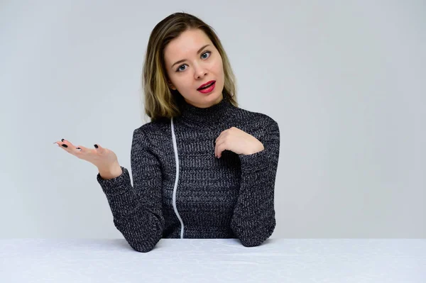 Concepto de una mujer parlante sentada en una mesa frente a la cámara. Foto de una chica bastante sonriente con el pelo rizado con emociones en un vestido gris sobre un fondo blanco . — Foto de Stock