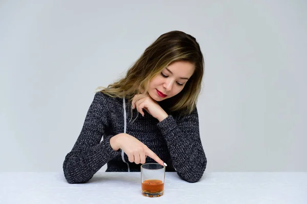 Retrato de una mujer sentada en la mesa hablando con el pelo rizado con emociones en un vestido gris sobre un fondo blanco con un vaso de whisky en las manos. Concepto de alcoholismo femenino . — Foto de Stock