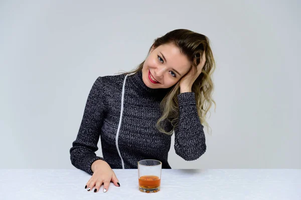 Retrato de una mujer sentada en la mesa hablando con el pelo rizado con emociones en un vestido gris sobre un fondo blanco con un vaso de whisky en las manos. Concepto de alcoholismo femenino . — Foto de Stock