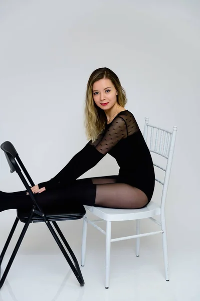 Art Concept van een modieus sexy meisje zittend op een stoel voor de camera. Foto van een glamoureuze mooie lachende vrouw met krullend haar in een sexy bodysuit op een witte achtergrond. — Stockfoto