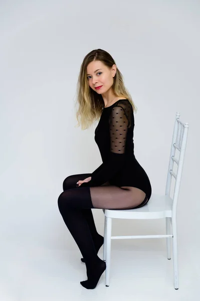Conceito de arte de uma menina sexy na moda sentada em uma cadeira na frente da câmera. Foto de uma mulher muito sorridente glamourosa com cabelo encaracolado em um bodysuit sexy em um fundo branco . — Fotografia de Stock