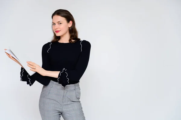 Concepto niña adulta sobre un fondo blanco con una carpeta blanca en las manos. Una foto de una guapa morena en pantalones grises y un suéter negro sonríe y muestra diferentes emociones en diferentes poses . — Foto de Stock