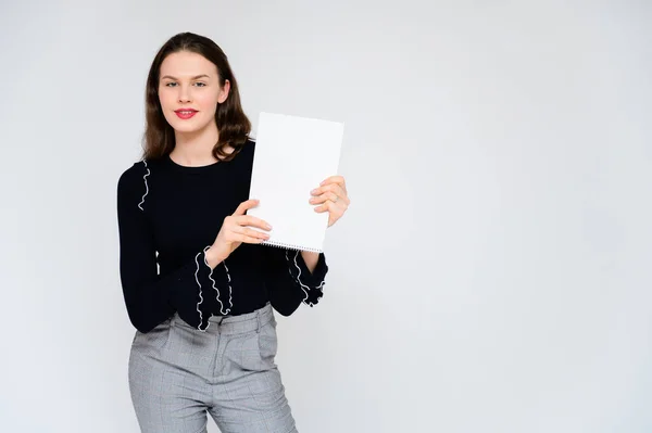 Concepto niña adulta sobre un fondo blanco con una carpeta blanca en las manos. Una foto de una guapa morena en pantalones grises y un suéter negro sonríe y muestra diferentes emociones en diferentes poses . — Foto de Stock