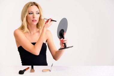 Güzellik ve kozmetik konsepti. Mükemmel makyajlı, güzel saçlı, beyaz arka planda temiz tenli güzel sarışın bir kadının portresi elinde bir aynayla makyaj yapıyor..