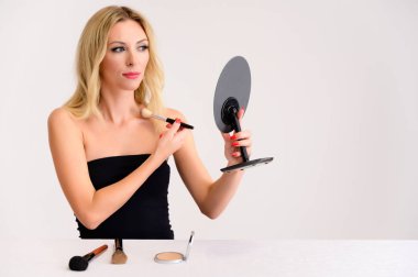 Güzellik ve kozmetik konsepti. Mükemmel makyajlı, güzel saçlı, beyaz arka planda temiz tenli güzel sarışın bir kadının portresi elinde bir aynayla makyaj yapıyor..