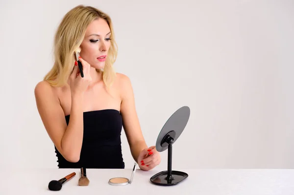 Beauty and cosmetics concept. Portrait of a pretty blonde model woman with excellent makeup, beautiful hair and clean skin on a white background is applying makeup with a mirror in her hands. — Stock Photo, Image