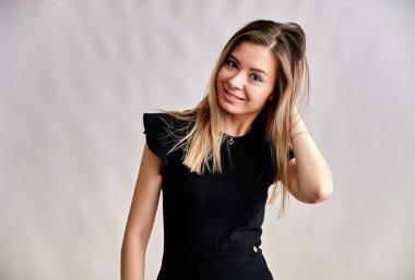 Gri arka planda, uzun düz saçlı, siyah elbiseli, gülümseyen güzel bir kadının yakın plan portresi. Kameranın önünde durmak, duygularını gösterir, farklı pozlarda konuşur..