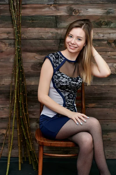 Portret ładnej uśmiechniętej dziewczyny z długimi włosami w szarej sukience na tle wnętrza w pokoju z drewna. Koncepcja stylu Loft. Siedzi na krześle, pokazuje słodkie szczęście. — Zdjęcie stockowe