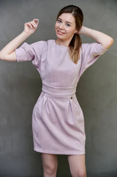 Retrato de una linda chica bastante sonriente en un vestido rosa con el pelo largo sobre un fondo de arte gris. Se mantiene derecho, muestra emociones, dulce felicidad . — Foto de Stock