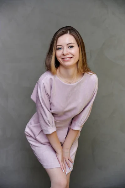 Retrato de uma menina bonita e sorridente em um vestido rosa com cabelos longos em um fundo de arte cinza. Fica em linha reta, mostra emoções, doce felicidade . — Fotografia de Stock