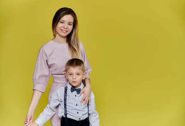 Portrait of a happy family: mom and a cute boy of 10 years of schoolboy on a yellow background in trousers and a shirt. Standing right in front of the camera, showing emotions, smile — Stock Photo, Image