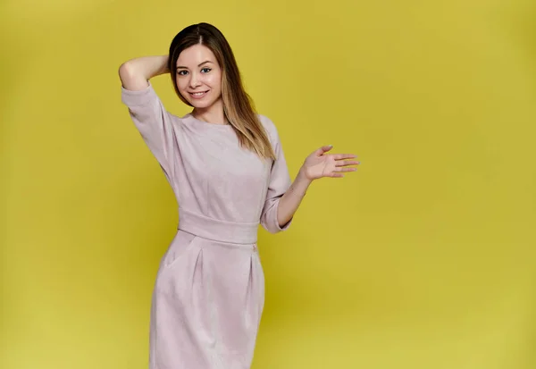Retrato de uma mulher muito jovem e sorridente em um fundo amarelo em um vestido rosa com cabelos longos e lisos. De pé bem na frente da câmera, mostra emoções, fala em diferentes poses . — Fotografia de Stock