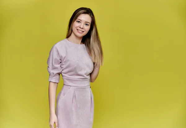 Retrato de uma mulher muito jovem e sorridente em um fundo amarelo em um vestido rosa com cabelos longos e lisos. De pé bem na frente da câmera, mostra emoções, fala em diferentes poses . — Fotografia de Stock