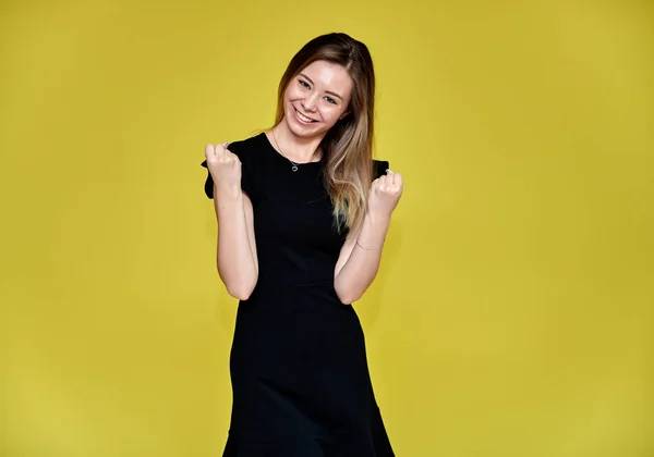 Retrato de uma mulher muito jovem e sorridente em um fundo amarelo em um vestido preto com cabelos longos e lisos. De pé bem na frente da câmera, mostra emoções, fala em diferentes poses . — Fotografia de Stock
