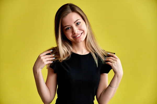 Retrato de cerca de una hermosa joven sonriente sobre un fondo amarillo en un vestido negro con el pelo largo y liso. De pie frente a la cámara, muestra emociones, habla en diferentes poses . — Foto de Stock