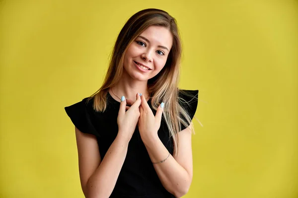 Retrato de cerca de una hermosa joven sonriente sobre un fondo amarillo en un vestido negro con el pelo largo y liso. De pie frente a la cámara, muestra emociones, habla en diferentes poses . — Foto de Stock