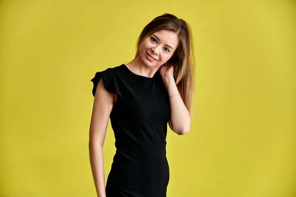 Retrato de uma menina muito jovem sorridente em um fundo amarelo em um vestido preto com cabelos longos e lisos. De pé bem na frente da câmera, mostra emoções, fala em diferentes poses . — Fotografia de Stock