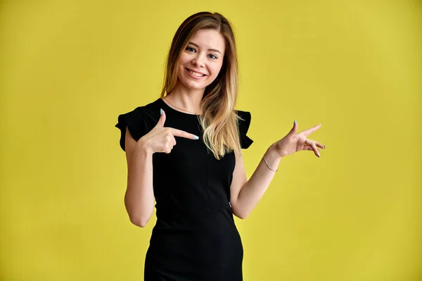Retrato de uma menina muito jovem sorridente em um fundo amarelo em um vestido preto com cabelos longos e lisos. De pé bem na frente da câmera, mostra emoções, fala em diferentes poses . — Fotografia de Stock