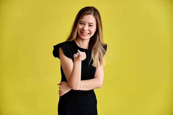 Retrato de una bonita joven sonriente sobre un fondo amarillo en un vestido negro con pelo largo y liso. De pie frente a la cámara, muestra emociones, habla en diferentes poses . — Foto de Stock