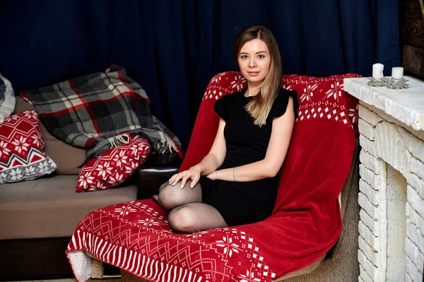 Retrato de una hermosa joven con el pelo largo en un vestido negro sentado en casa en un sofá en un estilo loft. Sentada frente a la cámara, mostrando emociones, sonriendo — Foto de Stock