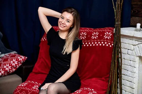 Porträt einer hübschen jungen Frau mit langen Haaren in einem schwarzen Kleid, die zu Hause auf einem Sofa im Loft-Stil sitzt. direkt vor der Kamera sitzen, Emotionen zeigen, lächeln — Stockfoto