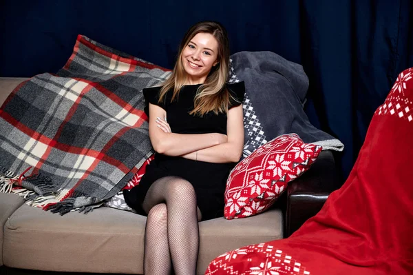 Retrato de una hermosa chica con el pelo largo en un vestido negro sentado en casa en un sofá en un estilo loft. Sentada frente a la cámara, mostrando emociones, sonriendo — Foto de Stock