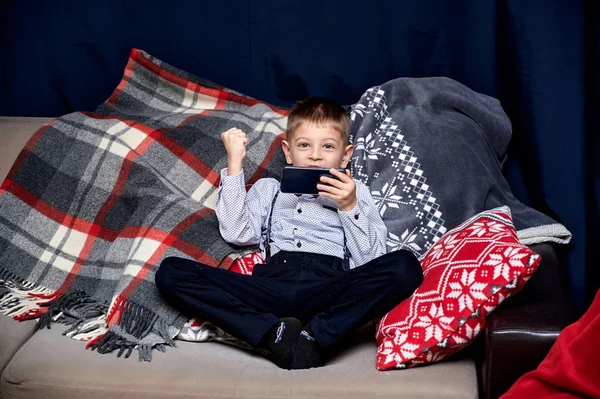 Portrait of a cute boy 10 year old schoolboy with a smartphone plays games at home on the sofa. Sitting right in front of the camera, showing emotions, smile — Stock Photo, Image