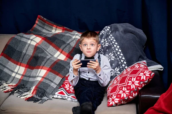 Portret van een leuke jongen 10 jaar oude schooljongen met een smartphone speelt games thuis op de bank. Recht voor de camera zitten, emoties tonen, glimlachen — Stockfoto