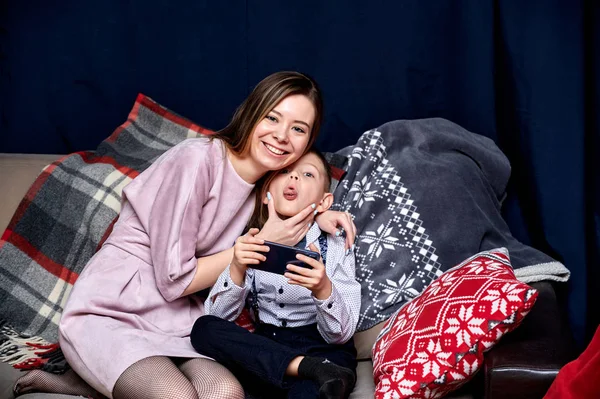 Портрет щасливої сім'ї: мама і милий хлопчик 10 років школярки зі смартфоном вдома на дивані. Сидячи прямо перед камерою, показуючи емоції, посміхаючись — стокове фото