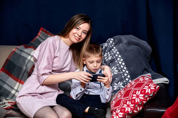 Portrait of a happy family: mom and a cute boy of 10 years of schoolboy with a smartphone at home on the sofa. Sitting right in front of the camera, showing emotions, smile — Stock Photo, Image