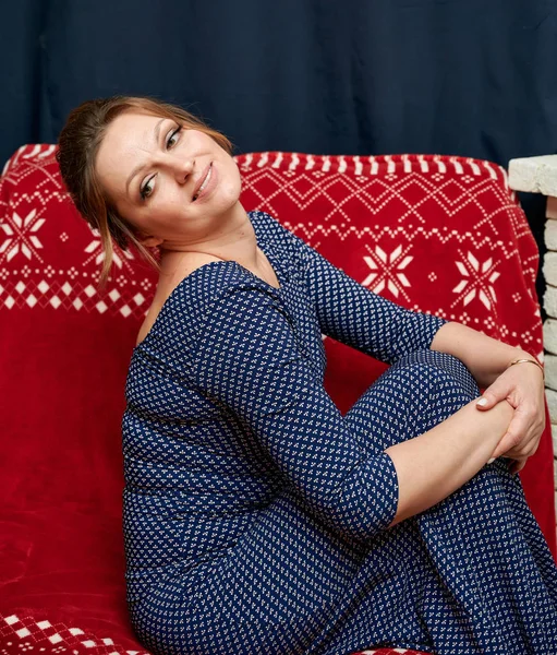 Retrato de una mujer adulta bonita de 40 años con buen maquillaje en un vestido largo en el interior de la casa cerca de la chimenea. Se sienta en un sofá justo delante de la cámara con una sonrisa . — Foto de Stock