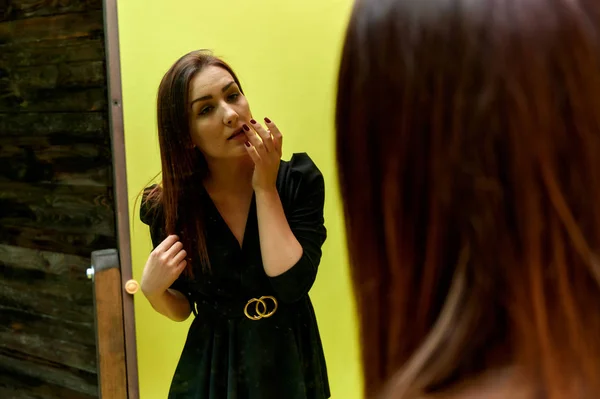 El concepto de un retrato glamoroso delante de un espejo. Retrato de una guapa morena vestida con un traje de moda negro revisando el maquillaje frente al espejo original . —  Fotos de Stock