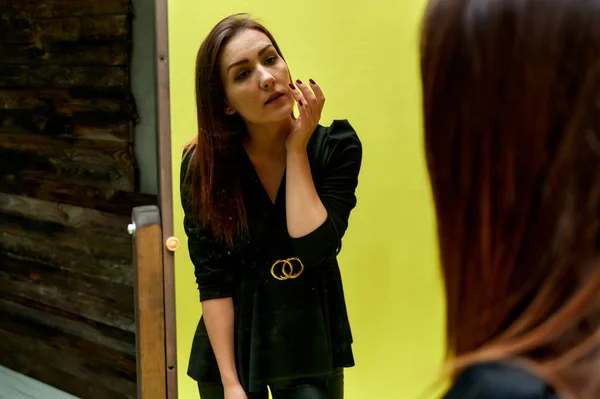 O conceito de um retrato glamouroso na frente de um espelho. Retrato de uma menina morena bonita em um terno de moda preta verificando maquiagem na frente do espelho original . — Fotografia de Stock