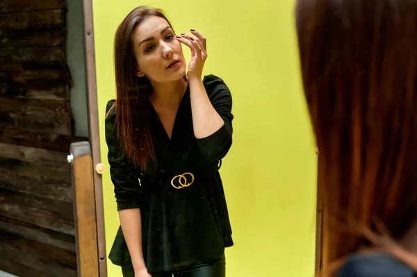 O conceito de um retrato glamouroso na frente de um espelho. Retrato de uma menina morena bonita em um terno de moda preta verificando maquiagem na frente do espelho original . — Fotografia de Stock