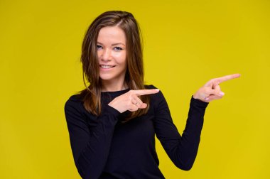 Olumlu duyguları olan gülümseyen tatlı bir kız konsepti. Siyah tişörtlü genç bir kadının portresi sarı arka planda uzun saçlı farklı pozlarda duruyor. Elleri göster, konuş..