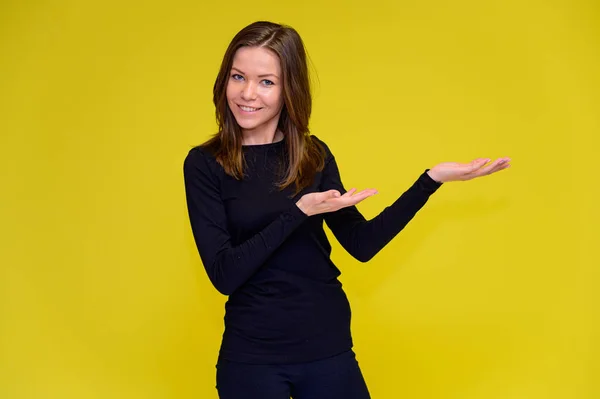 El concepto de una linda chica sonriente con emociones positivas. Retrato de una joven con una camiseta negra en diferentes poses con el pelo largo sobre un fondo amarillo. Muestra las manos, habla . — Foto de Stock