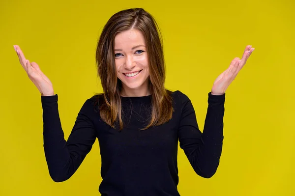 El concepto de una linda chica sonriente con emociones positivas. Retrato de una joven con una camiseta negra en diferentes poses con el pelo largo sobre un fondo amarillo. Muestra las manos, habla . — Foto de Stock