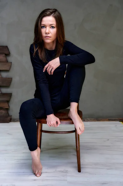Pionowe zdjęcie na krześle w stylowym wnętrzu w studiu dość efektownej brunetki. dziewczyna siedzi na krześle w czarny T-shirt z długimi włosami na modnym oryginalnym tle. — Zdjęcie stockowe