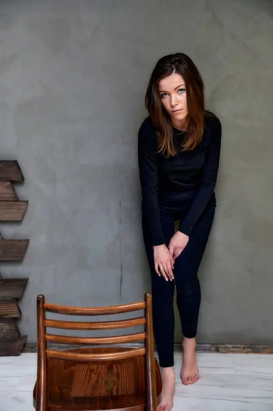 Pionowe zdjęcie z krzesłem w stylowym wnętrzu w studiu dość efektownej brunetki. dziewczyna stoi w czarny T-shirt z długimi włosami na alternatywnym kolorze modne oryginalne tło. — Zdjęcie stockowe