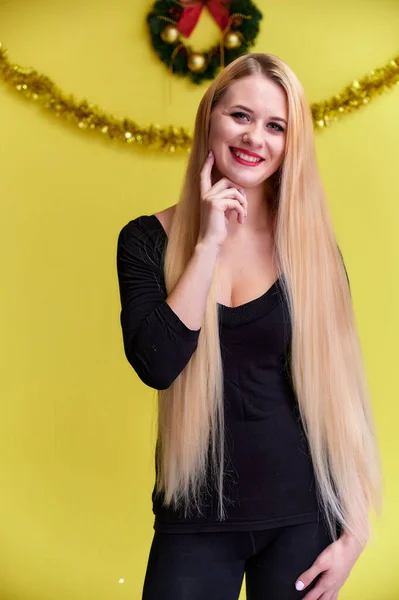 Konzept einer jungen blonden Frau mit Neujahrsdekor. Porträt eines süßen Mädchens in einem schwarzen T-Shirt mit langen, schönen Haaren und tollem Make-up. Lächeln, Emotionen auf gelbem Hintergrund zeigen. — Stockfoto