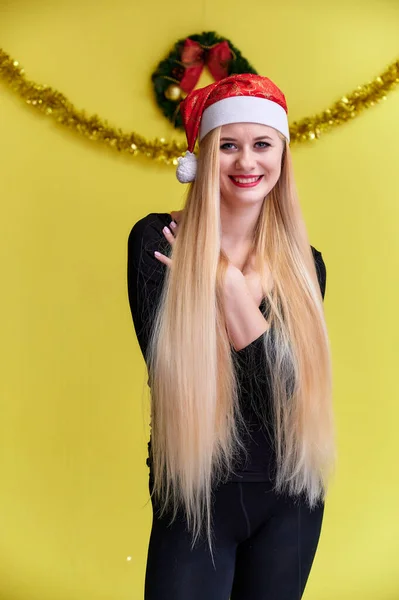 Koncept mladé blondýny s novoroční výzdobou. Portrét roztomilé dívky v černém tričku s dlouhými krásnými vlasy a skvělým make-upem. Usmívající se, ukazující emoce na žlutém pozadí. — Stock fotografie