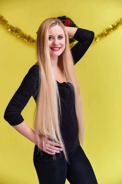 Έννοια μιας νεαρής ξανθιάς γυναίκας με πρωτοχρονιάτικη διακόσμηση. Πορτρέτο ενός χαριτωμένο κορίτσι σε ένα μαύρο T-shirt με μακριά όμορφα μαλλιά και μεγάλη μακιγιάζ. Χαμογελώντας, δείχνοντας συναισθήματα σε κίτρινο φόντο. — Φωτογραφία Αρχείου