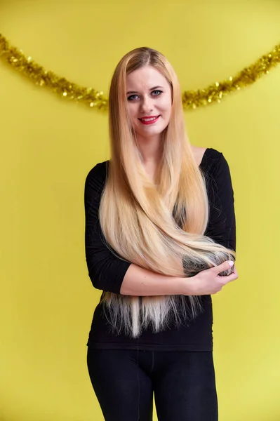 Concepto de una joven rubia con decoración de Año Nuevo. Retrato de una linda chica en una camiseta negra con pelo largo y hermoso y gran maquillaje. Sonriendo, mostrando emociones sobre un fondo amarillo . — Foto de Stock