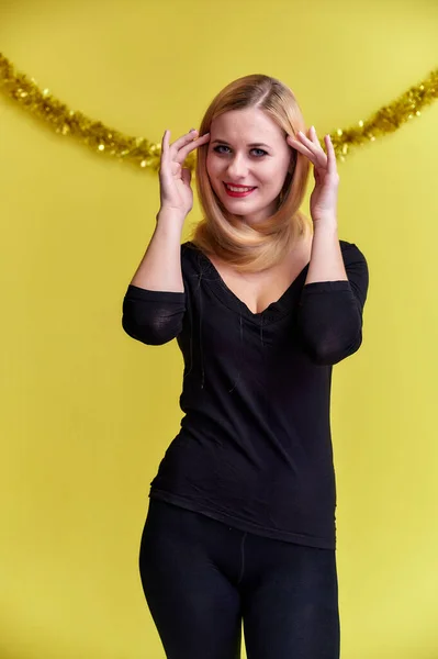 Koncept mladé blondýny s novoroční výzdobou. Portrét roztomilé dívky v černém tričku s dlouhými krásnými vlasy a skvělým make-upem. Usmívající se, ukazující emoce na žlutém pozadí. — Stock fotografie