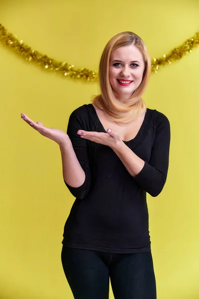 Concept van een jonge blonde vrouw met nieuwjaarsdecor. Portret van een schattig meisje in een zwart T-shirt met lang mooi haar en geweldige make-up. Glimlachen, emoties tonen op een gele achtergrond. — Stockfoto