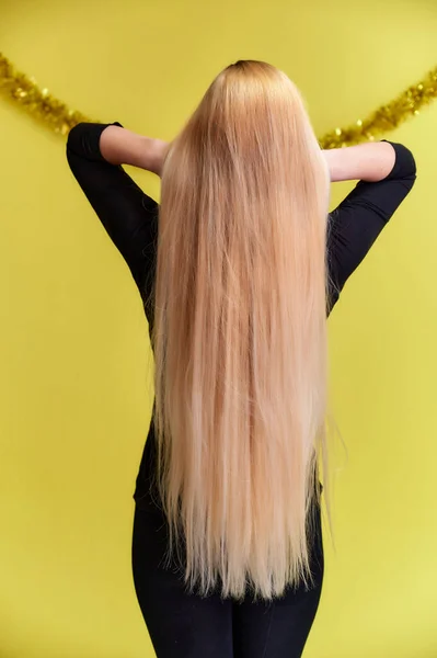 Concept of a young blonde woman with a Christmas decor, rear view. Portrait of a cute girl in a black T-shirt with long beautiful hair and great makeup. Smiling on a yellow background. — Stock Photo, Image