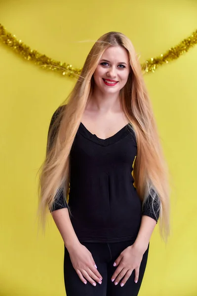 Koncepcja młodej blondynki z noworocznym wystrojem. Portret uroczej dziewczyny w czarnej koszulce z długimi, pięknymi włosami i świetnym makijażem. Uśmiechnięta, pokazująca emocje na żółtym tle. — Zdjęcie stockowe