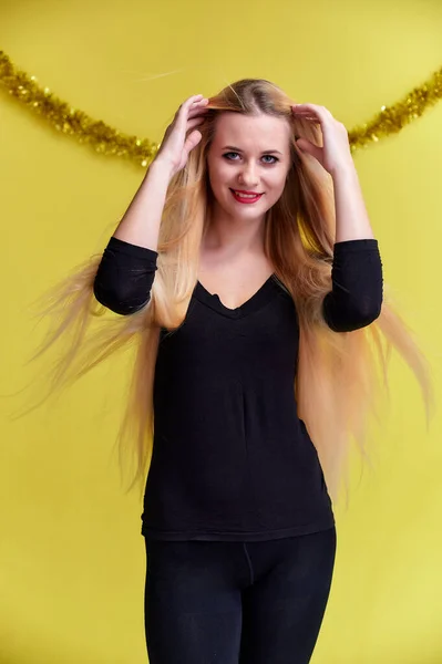 Concepto de una joven rubia con decoración de Año Nuevo. Retrato de una linda chica en una camiseta negra con pelo largo y hermoso y gran maquillaje. Sonriendo, mostrando emociones sobre un fondo amarillo . — Foto de Stock