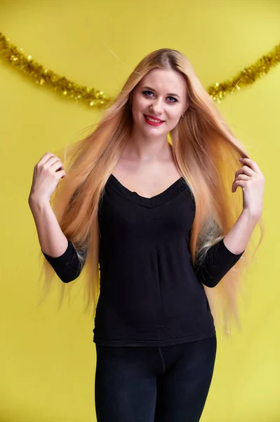 Concept van een jonge blonde vrouw met nieuwjaarsdecor. Portret van een schattig meisje in een zwart T-shirt met lang mooi haar en geweldige make-up. Glimlachen, emoties tonen op een gele achtergrond. — Stockfoto