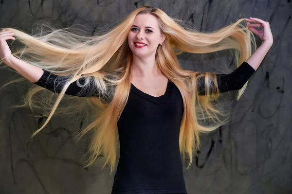 Concepto de una joven rubia con un peinado chic. Retrato de una linda chica en una camiseta negra con pelo largo y hermoso y gran maquillaje. Sonriendo, mostrando emociones sobre un fondo gris . — Foto de Stock